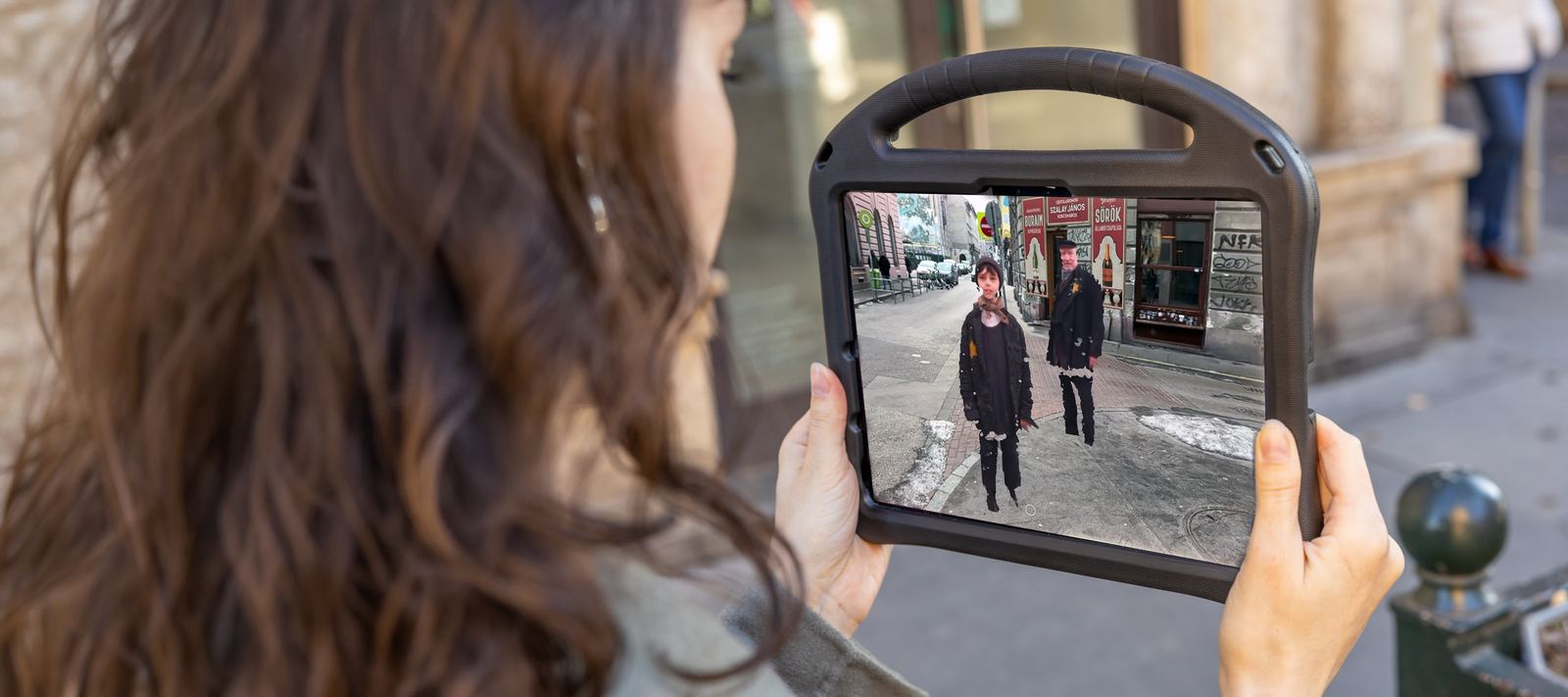 A visitor holds an iPad on a Budapest street. On the screen, two characters from the past stand in the same street.